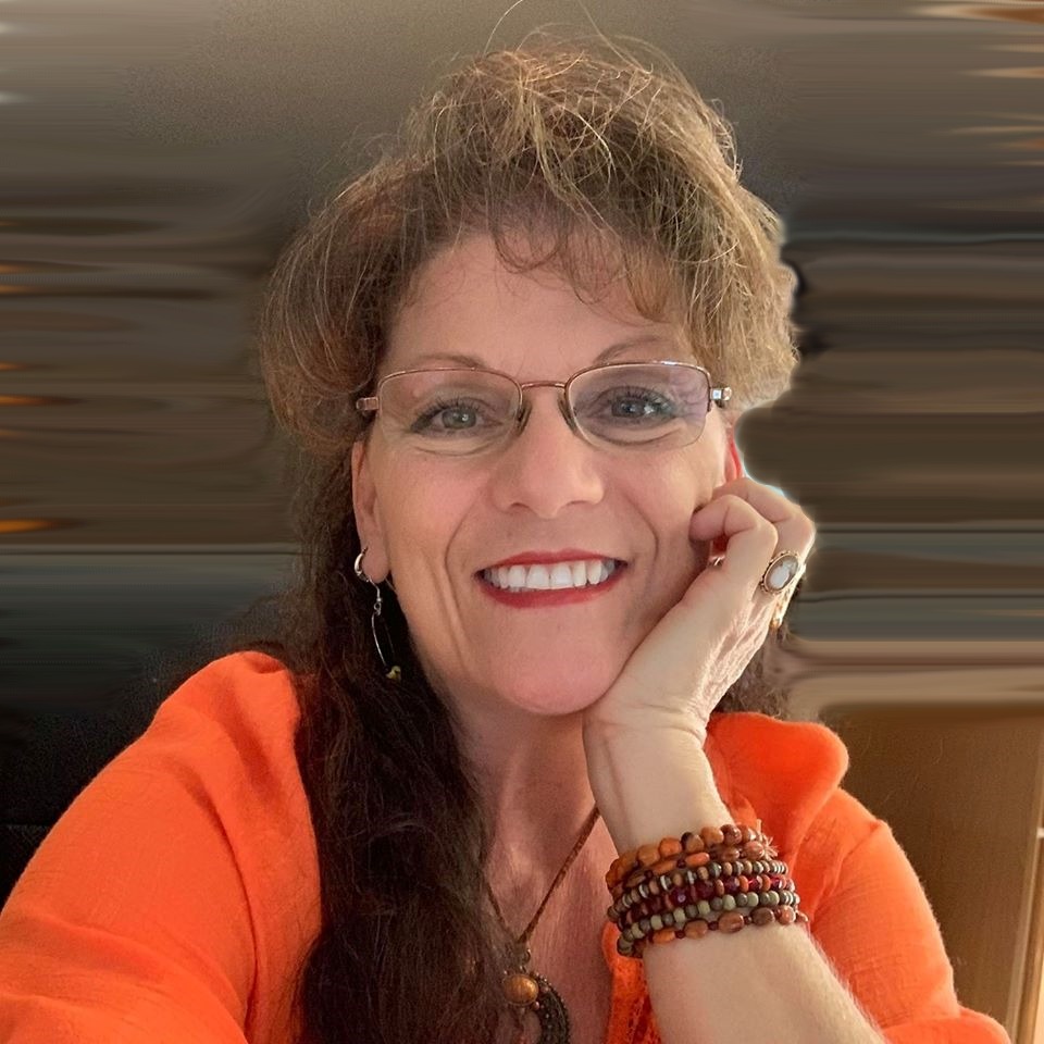 Smiling woman with glasses and curly hair, wearing an orange top, resting her face on her hand, with a blurred background.