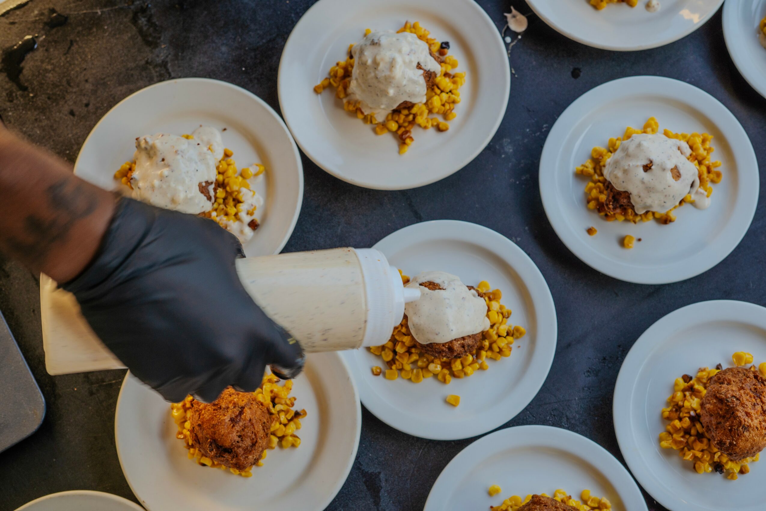 Person wearing a black glove serving food onto white plates with rice and fried items, on a dark surface.