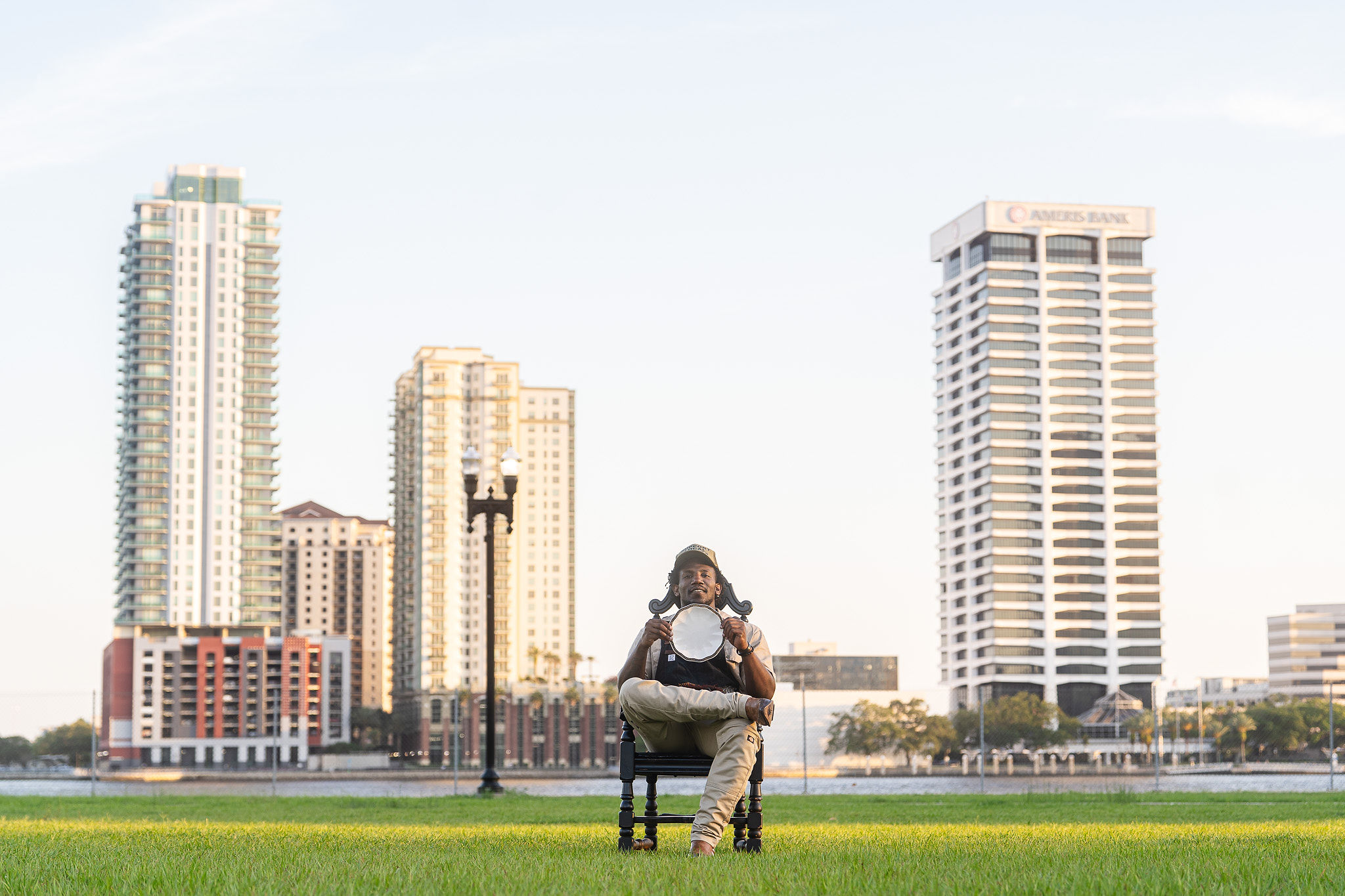 Person sitting on a bench facing a city skyline with tall buildings, water, and a grassy area in the foreground.