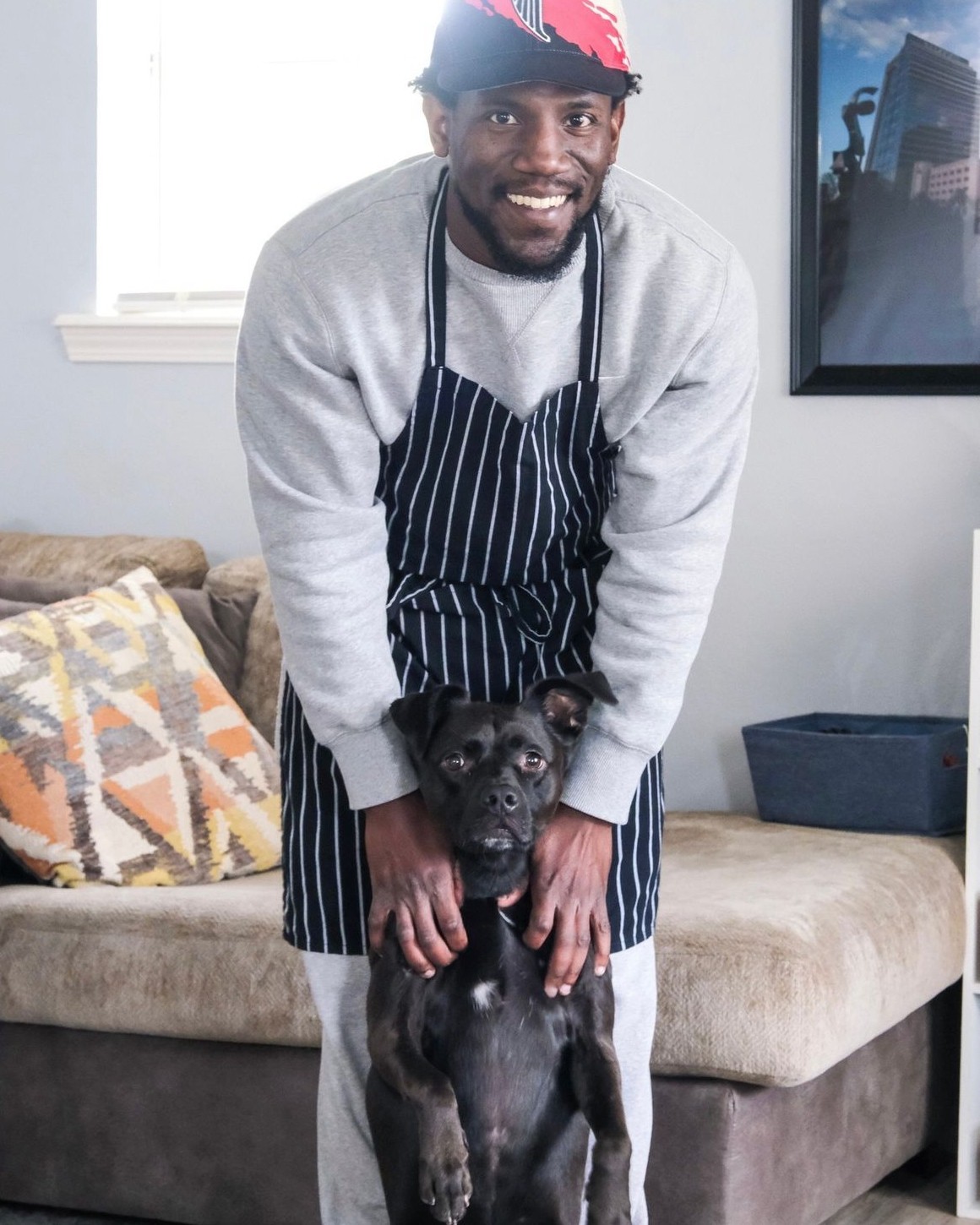 Man smiling, wearing apron, with a black dog in a living room, sofa with patterned pillow, TV on wall.