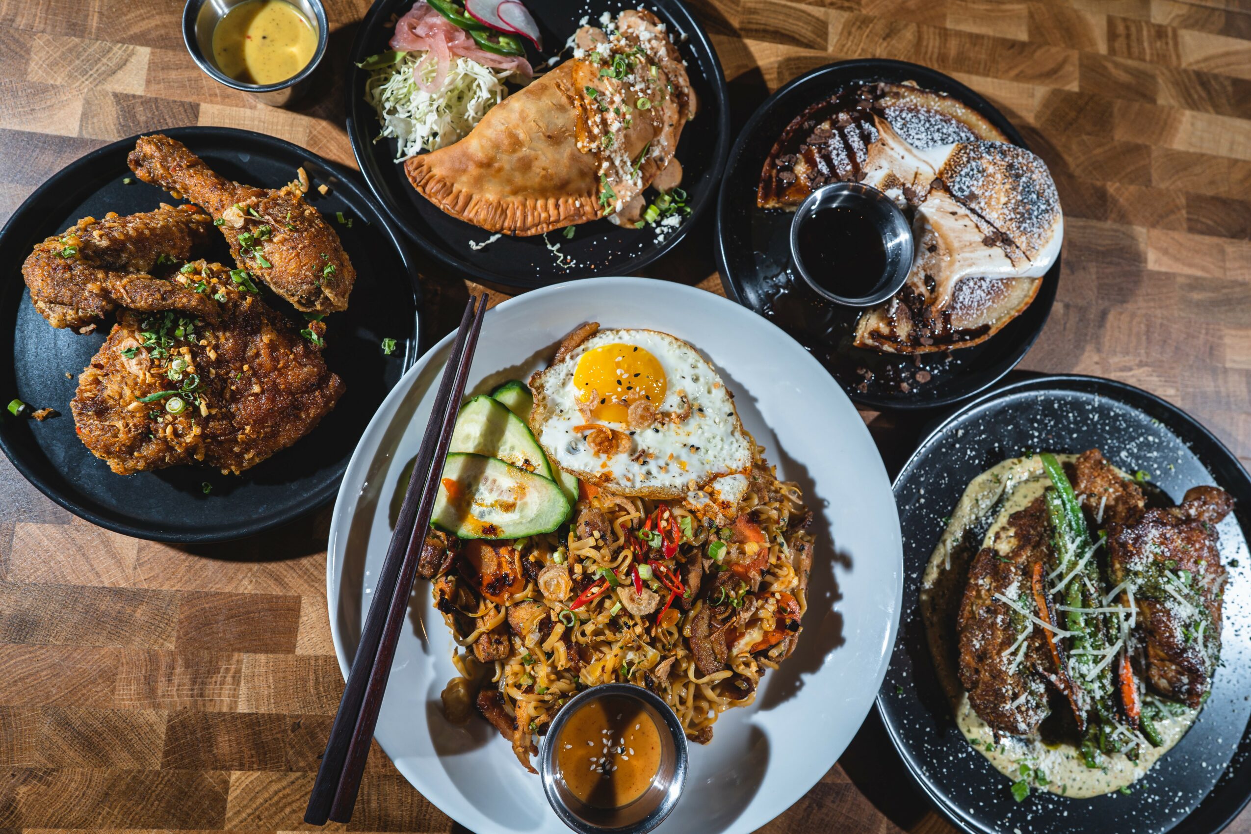 Assorted dishes including fried chicken, rice with vegetables and egg, samosas, burger, and drinks on a wooden table.