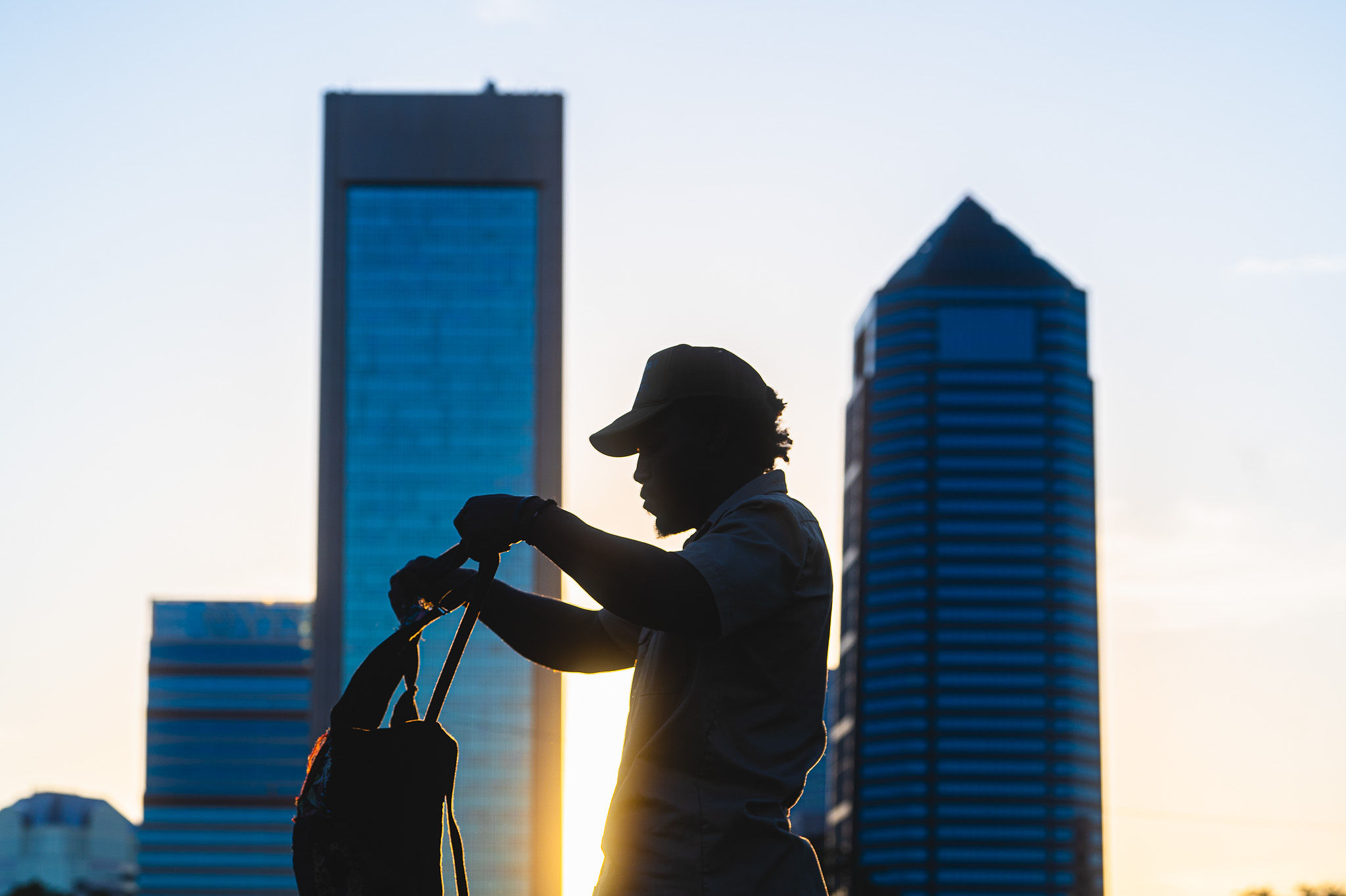 Silhouette of a person with a cap holding a bag against a city skyline at sunset.