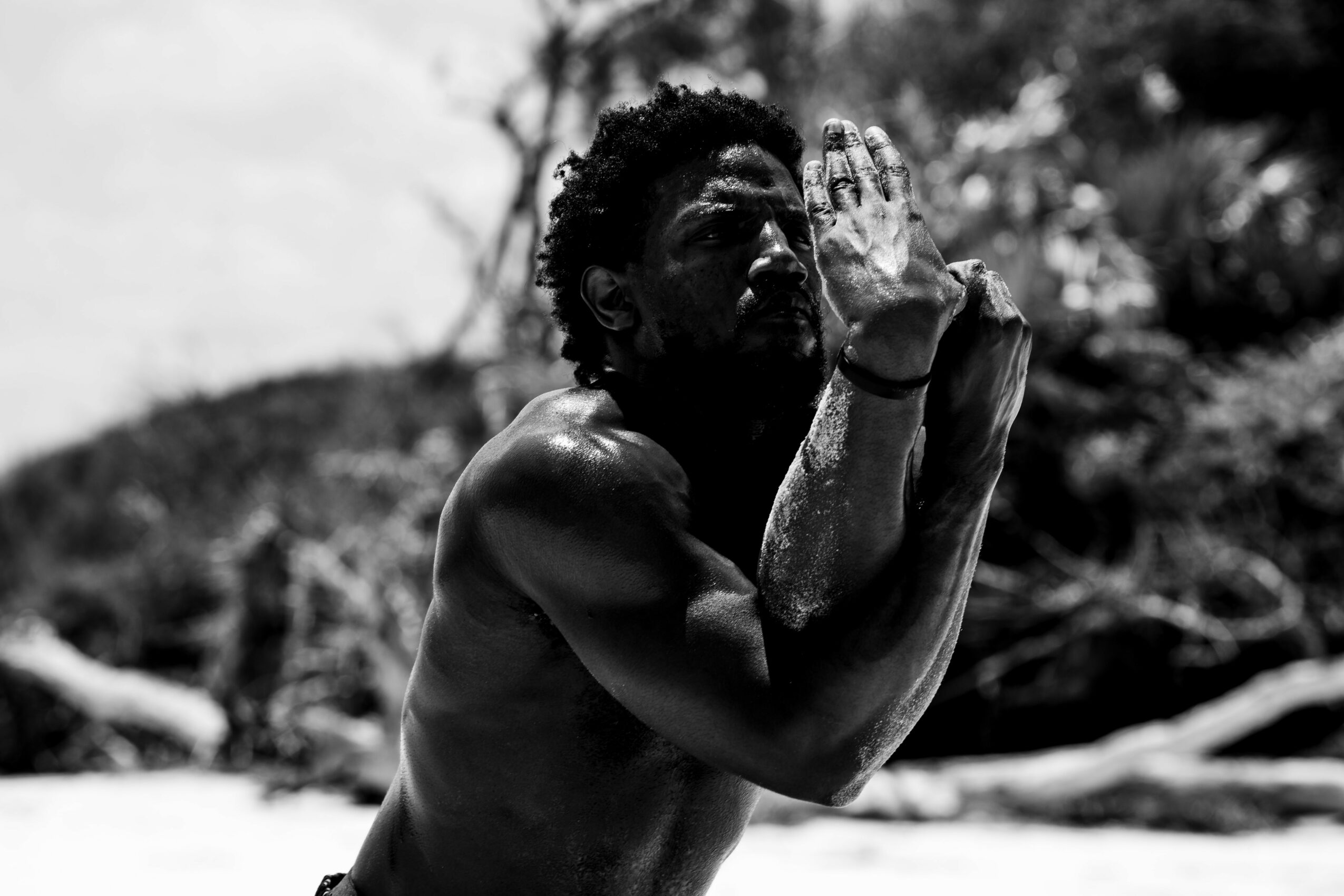 A shirtless man with curly hair holds his hands together near his face outdoors, with trees and sky in the background.