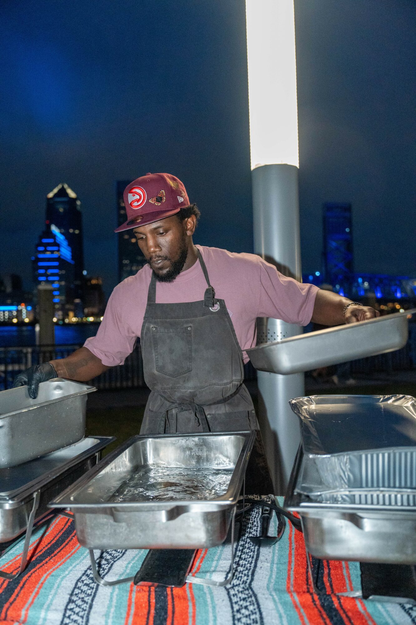 Person wearing a cap and apron preparing food outdoors at night with city skyline in background.