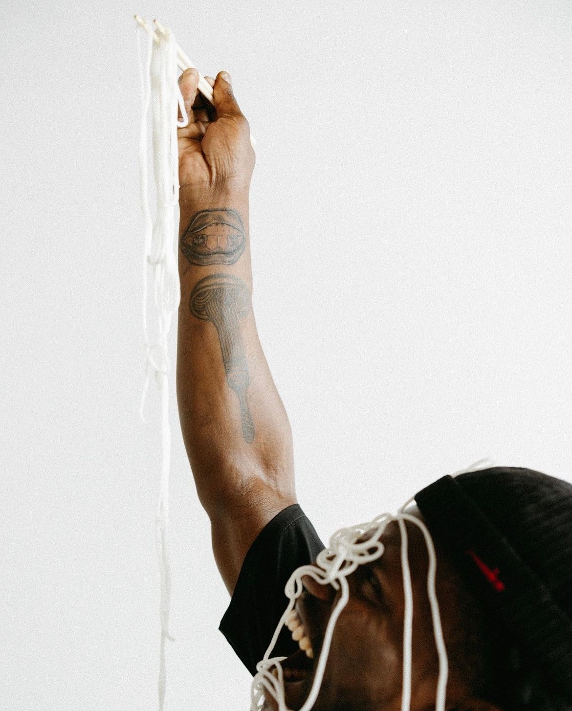 Person's arm with tattoos reaching upward, holding a white string or cord against a plain white background.