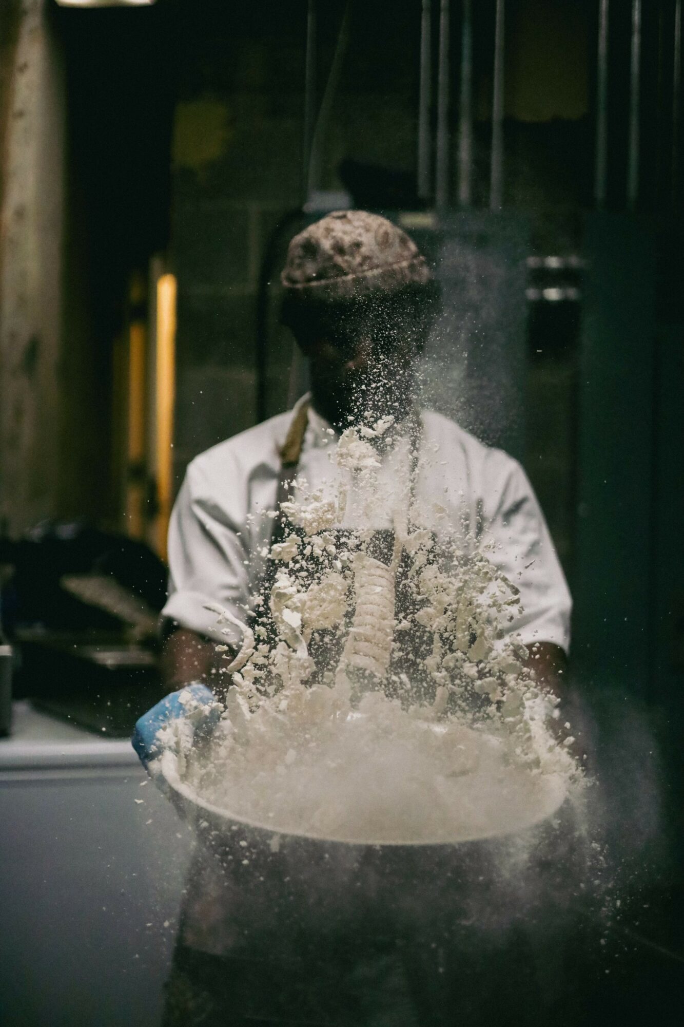 Person tossing dough in the air, flour flying, wearing a white shirt and apron, in a kitchen or bakery setting.