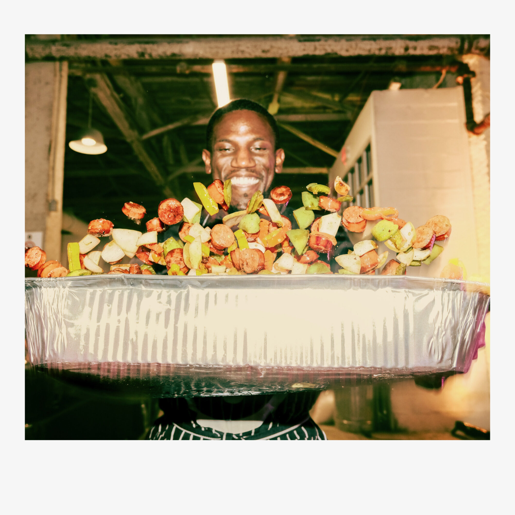 Person holding a large tray of colorful food, smiling, in an industrial setting with exposed beams and lighting.
