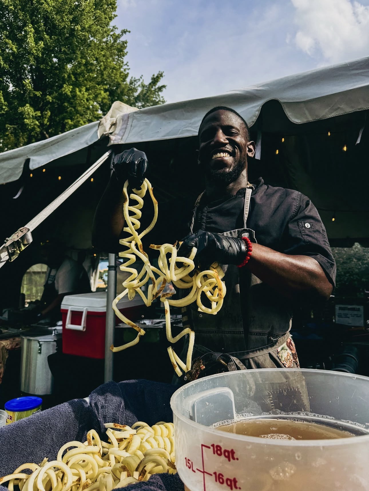 Man smiling holding a yellow noodle in an outdoor setting with a tent and trees in the background.