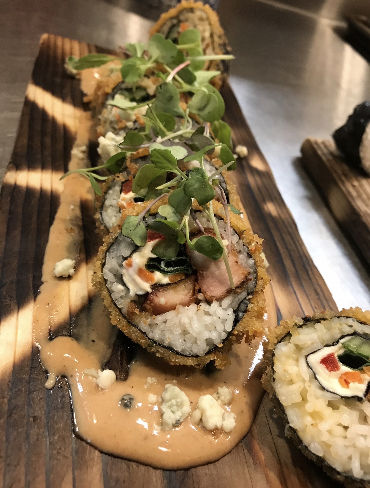Sushi rolls topped with microgreens and drizzled sauce on a wooden platter.