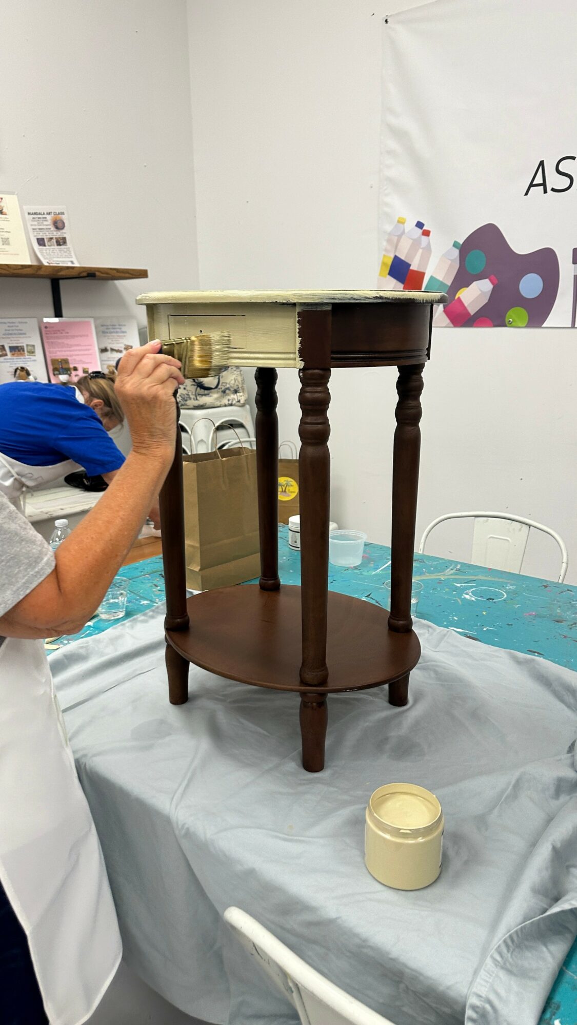 Person working on a small, ornate wooden table with a cream-colored top, in a room with a table and banner. Word 'AS' visible.