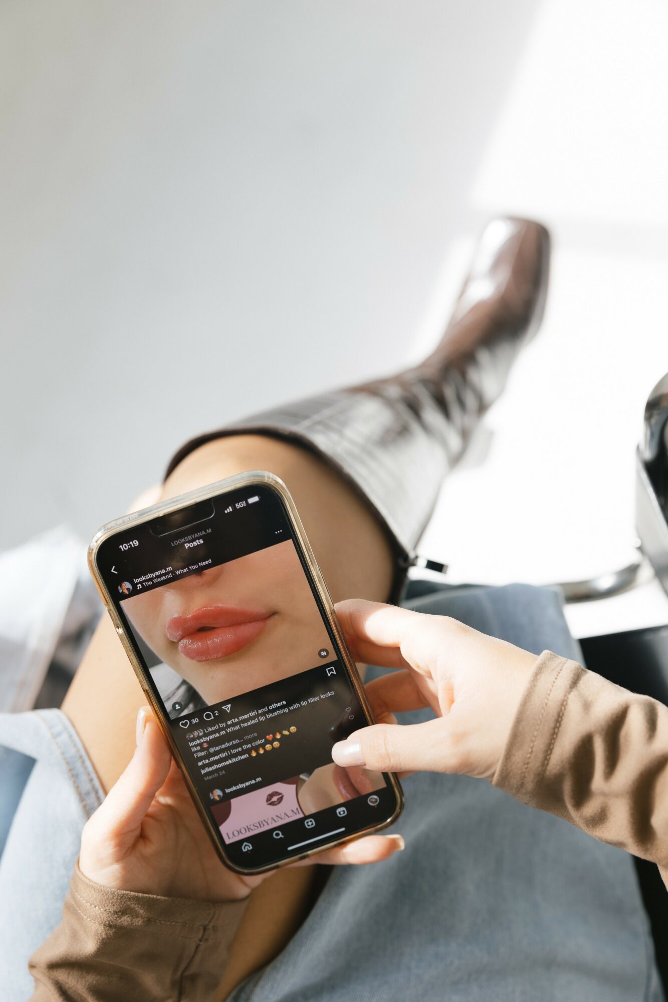 Person holding a smartphone capturing a close-up of lips, with legs crossed and a bright background.
