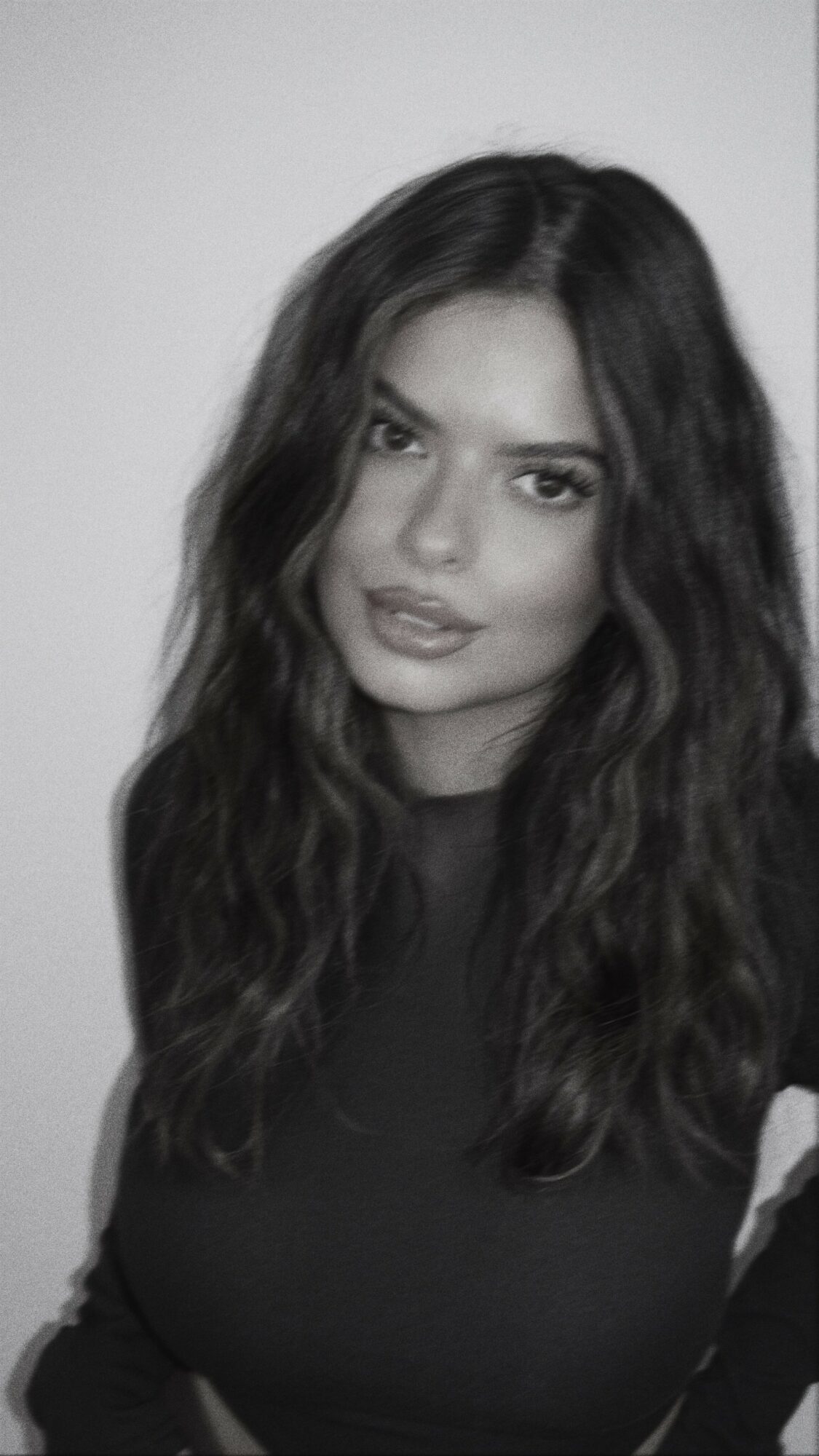 Young woman with long wavy hair looking at camera, wearing dark top, neutral background.