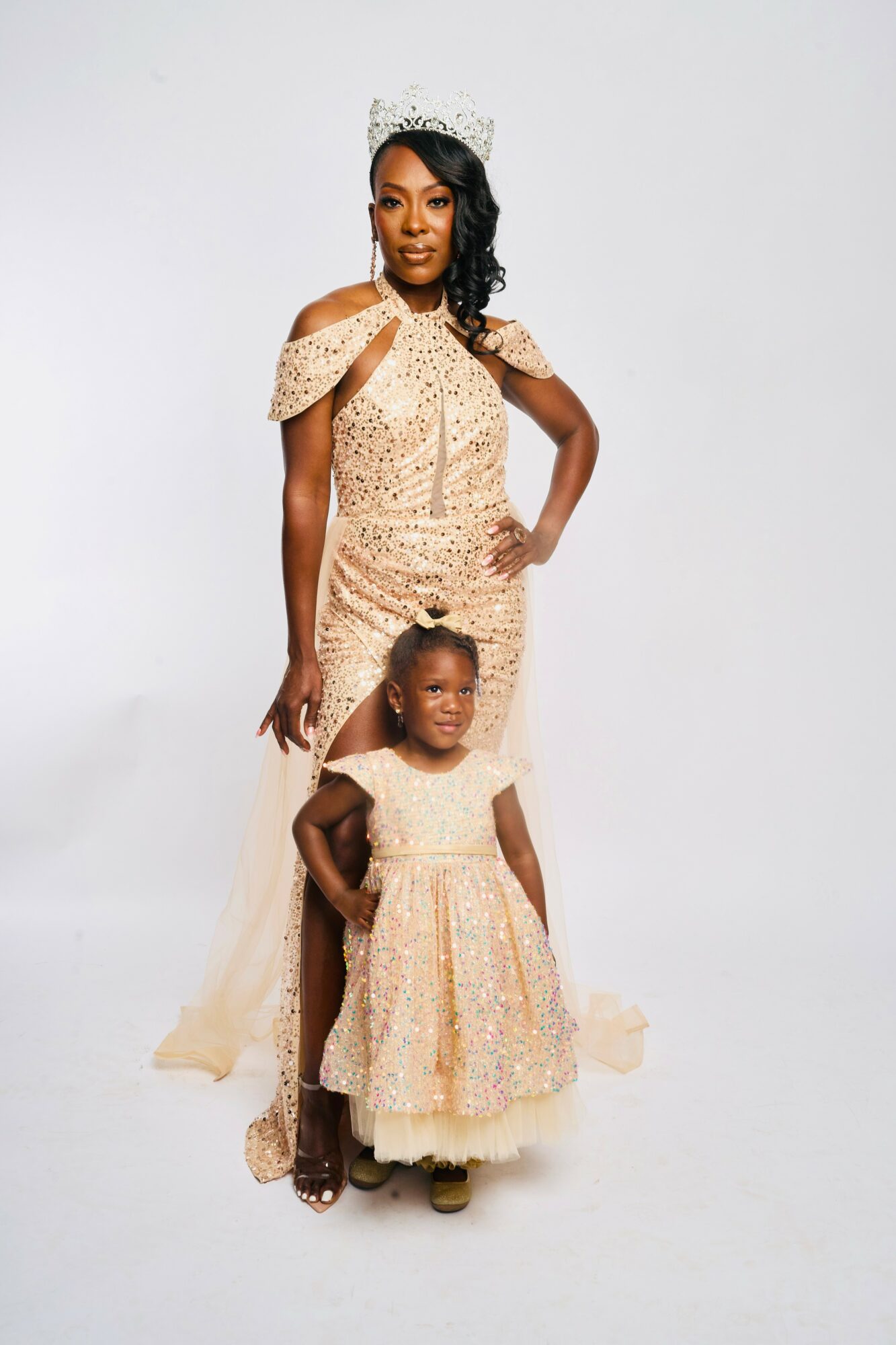 A woman and a young girl dressed in elegant, matching gold gowns with lace details, standing against a plain white background.