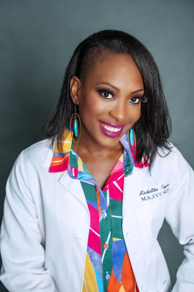 Smiling woman with dark hair, wearing colorful earrings and a white coat, against a plain background.
