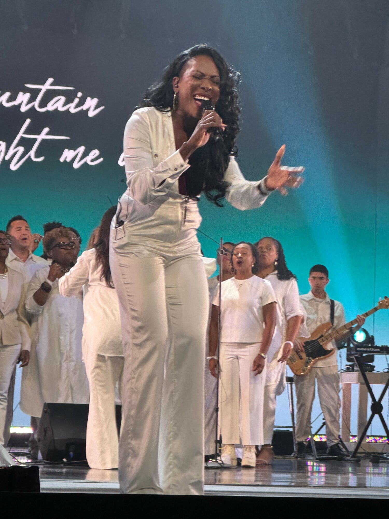 A woman in white pants and a white top speaking into a microphone on stage, with a group of people behind her.