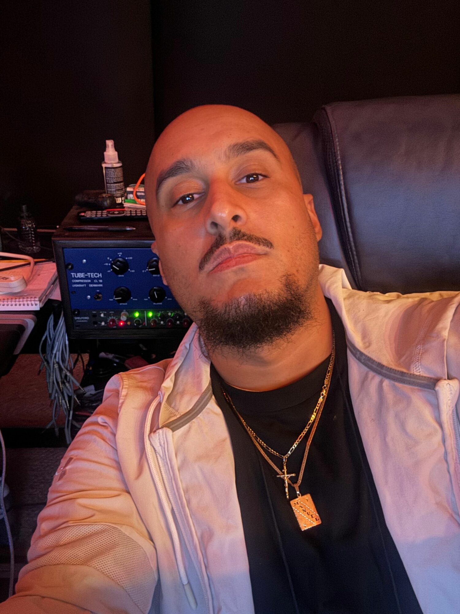 Man lying on a couch with a pillow, wearing a white jacket and gold chain, with electronic equipment nearby.
