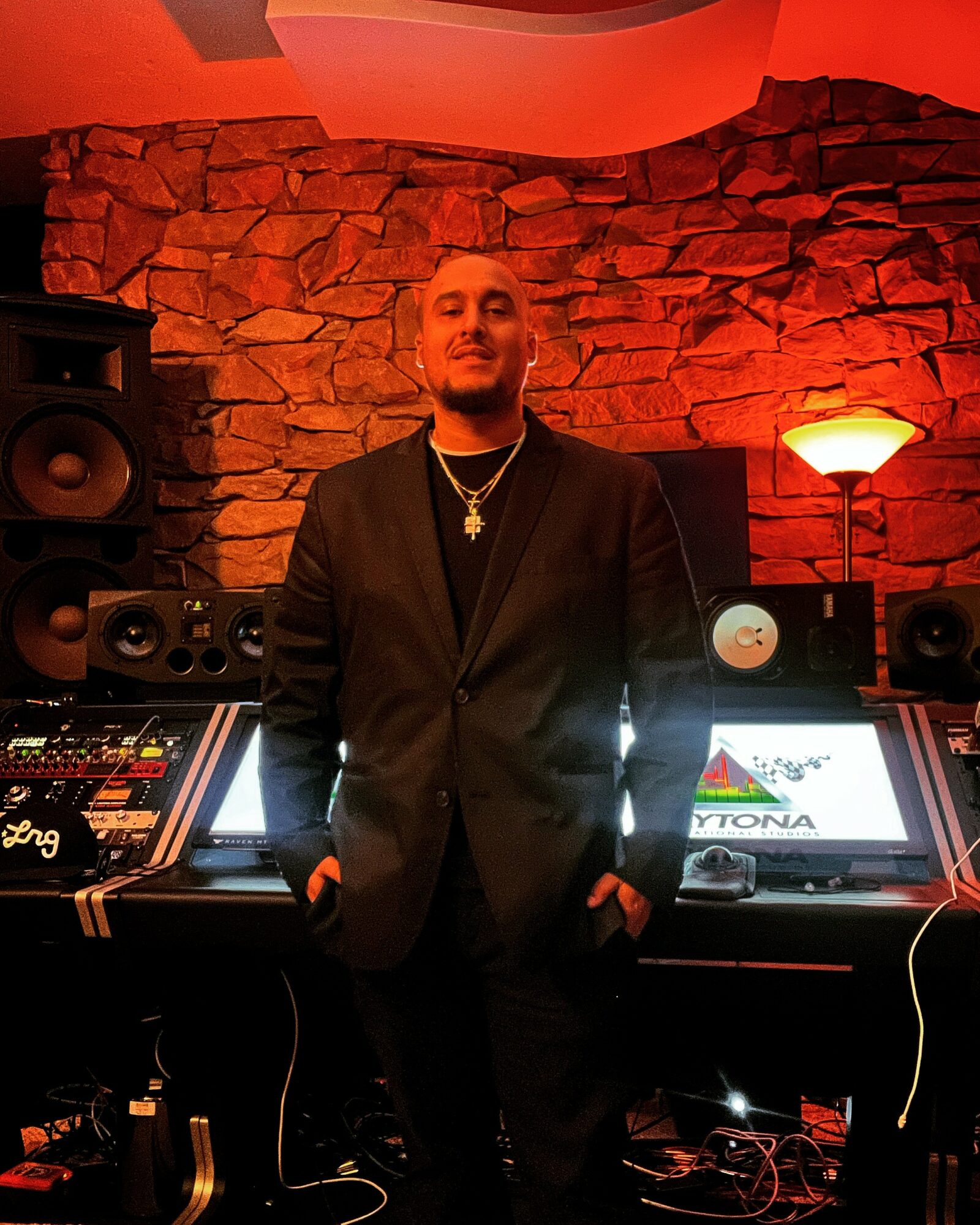 Man in black suit standing in a music studio with equipment and a stone wall background.