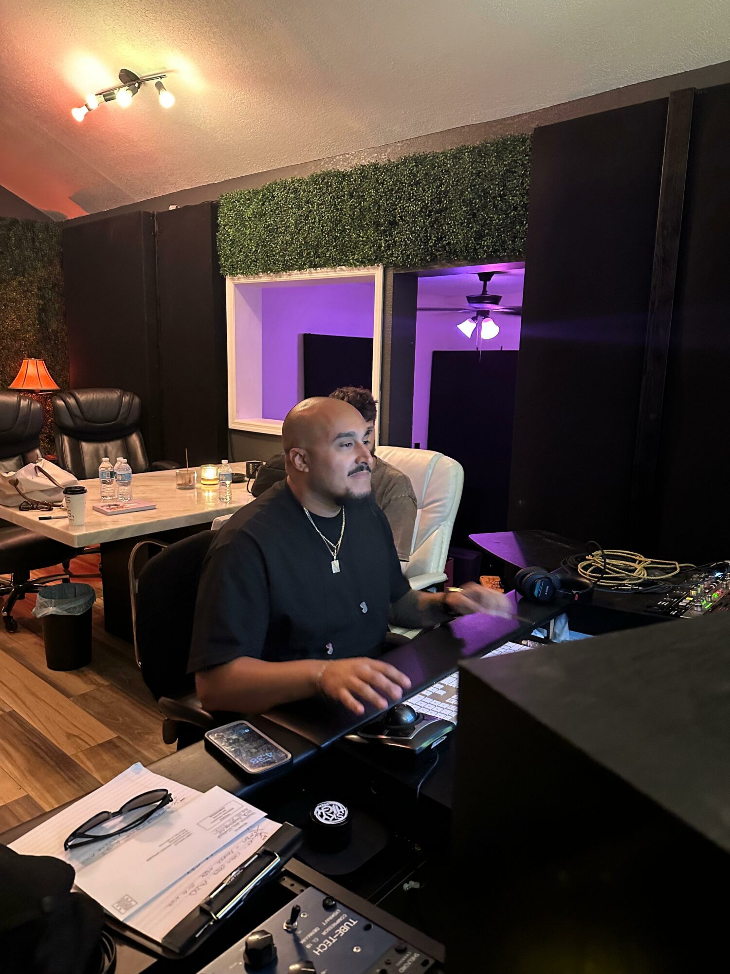 Person sitting at a desk in a room with purple lighting, black walls, and a green wall decoration, working on a computer.