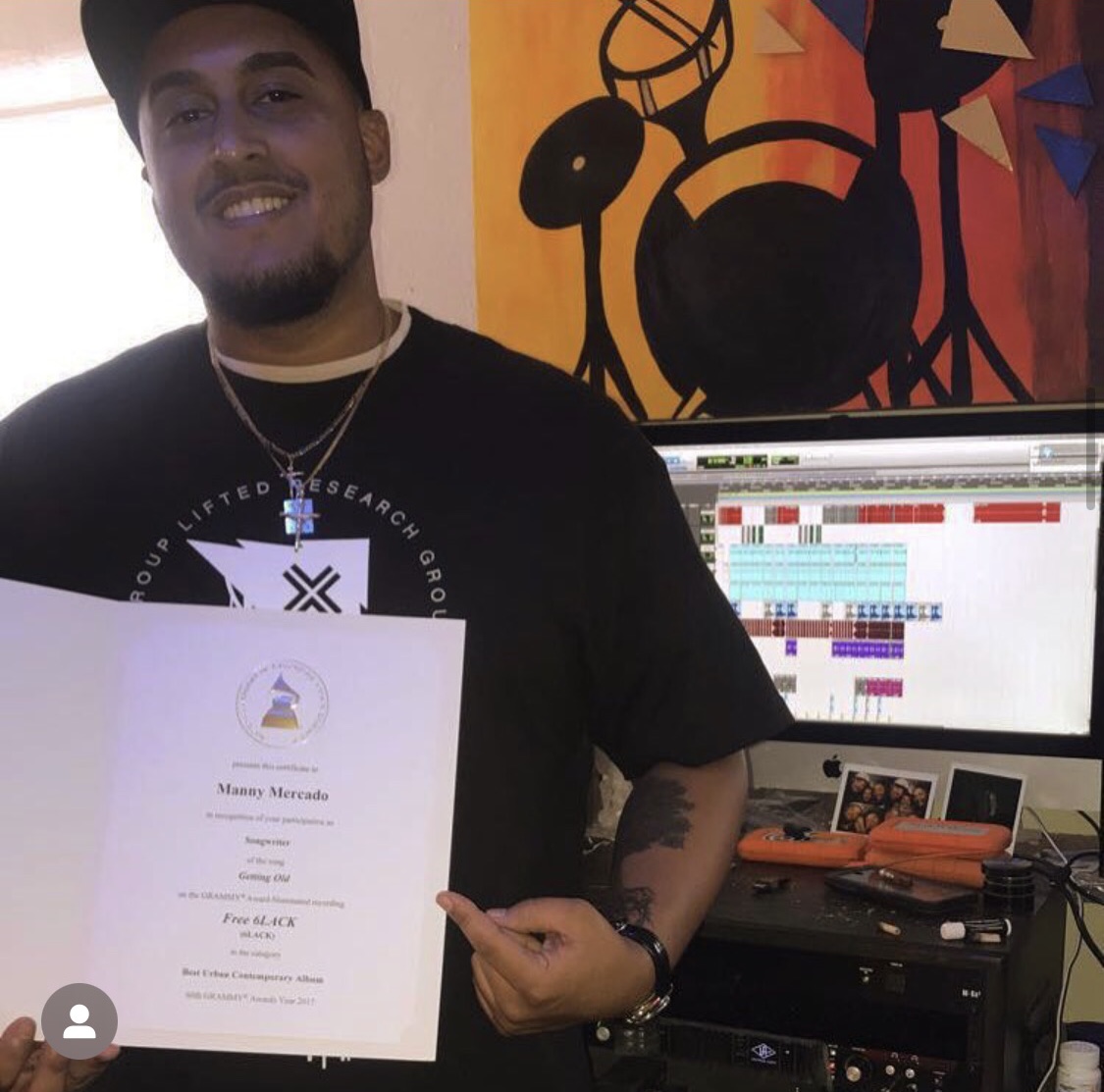Person holding a certificate, smiling, in a room with a colorful abstract painting and a computer monitor displaying a digital audio workstation.
