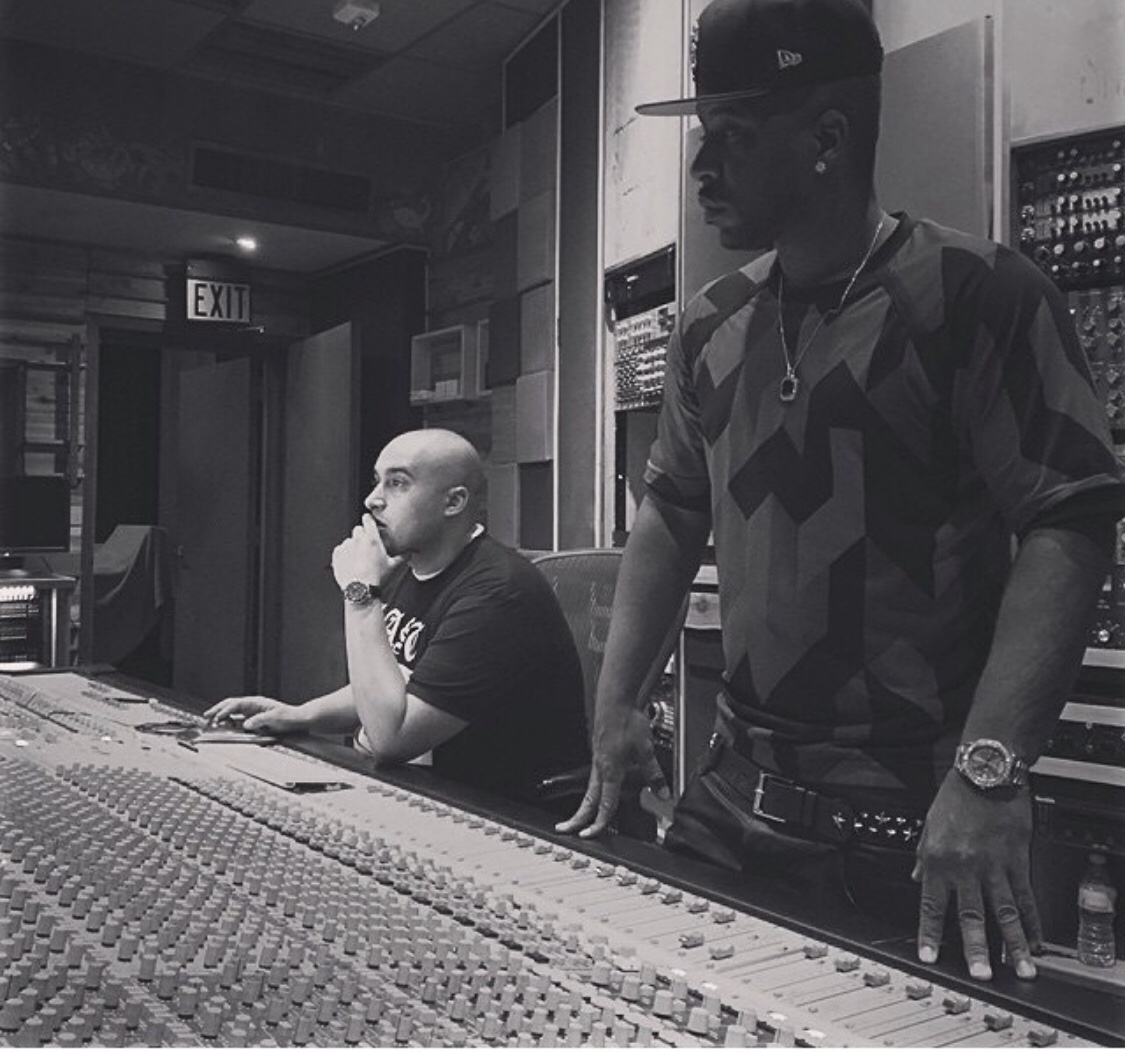 Two men in a recording studio, one sitting at a mixing console and the other standing nearby, both wearing casual clothing.