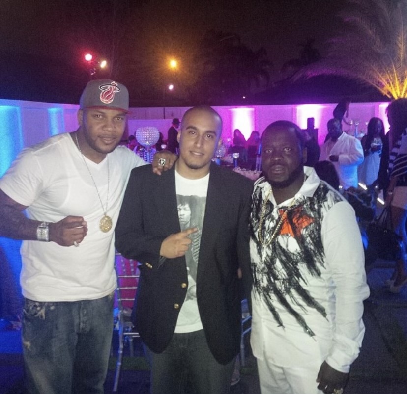 Three men standing together at a nighttime event with colorful lighting, smiling, with people in the background.