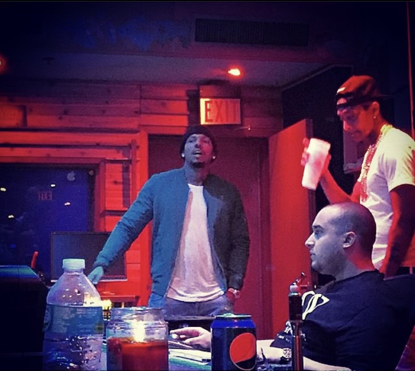 Three people in a room with red lighting, one standing, two seated, with drinks and bottles on the table.