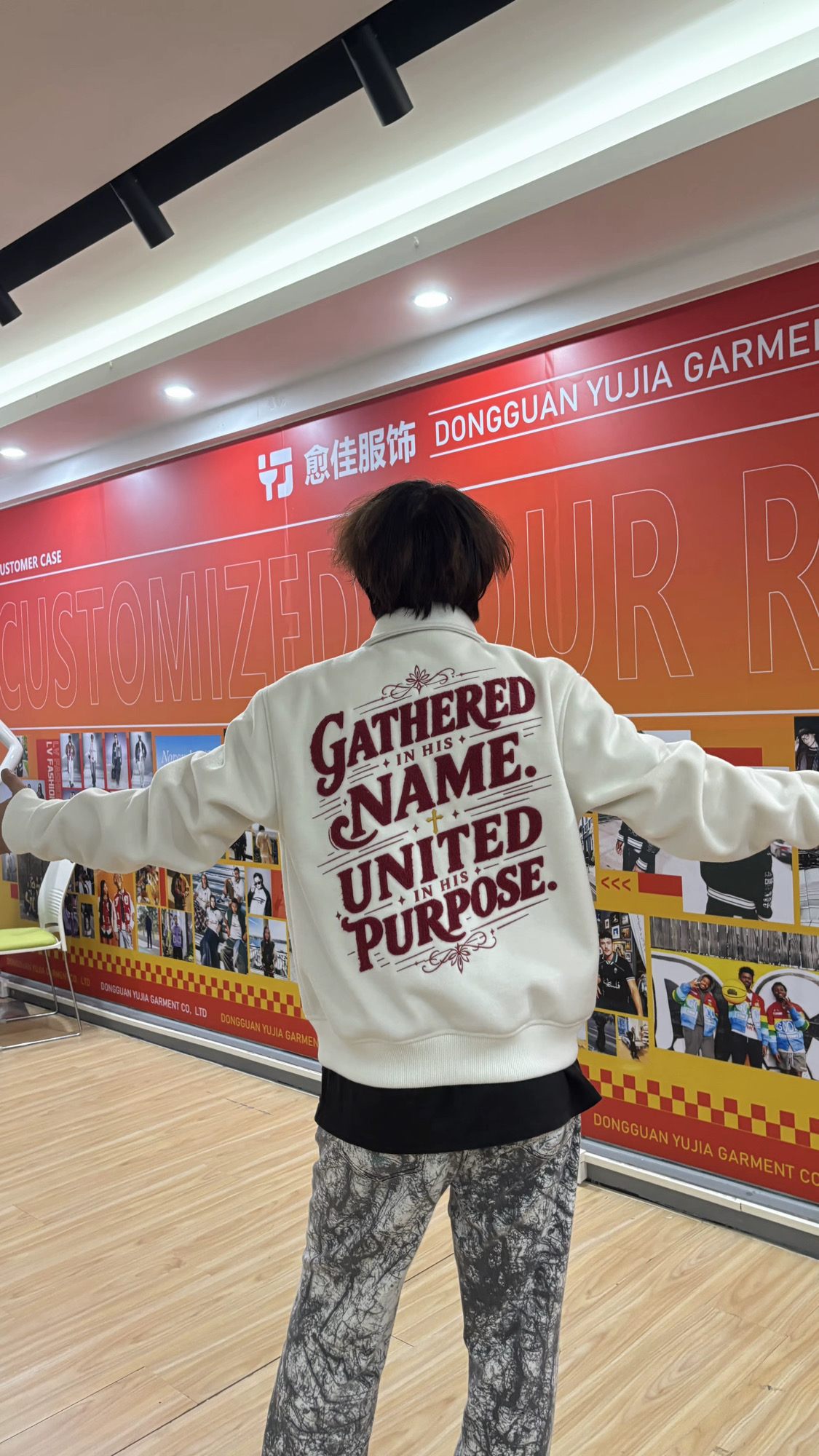 Person with arms outstretched wearing a white hoodie with red text, standing in front of a red wall with store signage.