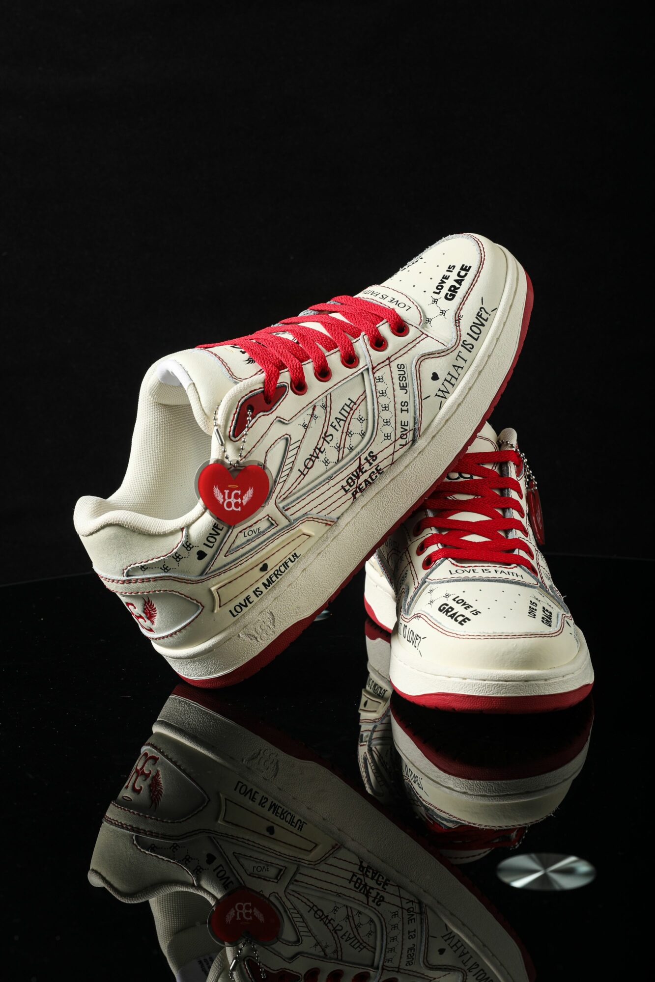 Pair of white sneakers with red laces and a heart-shaped tag, decorated with black text and patterns, on a reflective surface.
