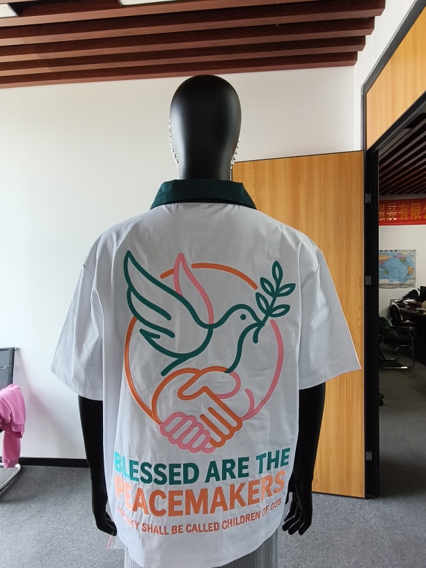 Black mannequin wearing a white T-shirt with a colorful dove, handshake, and text design, indoors with wooden and black elements.