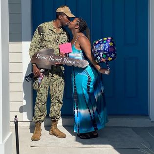 Person in military uniform and woman with a balloon and gift, sharing a kiss outside a house.