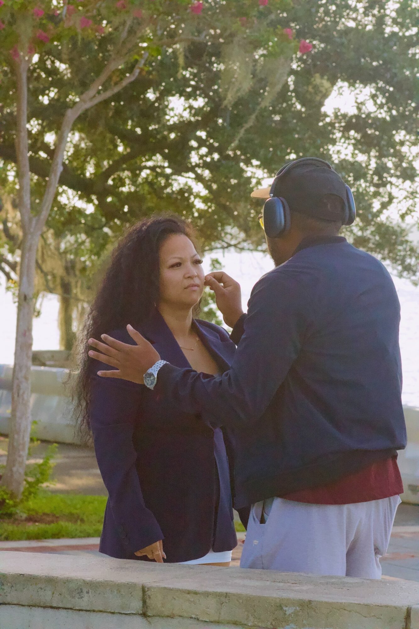 Two people outdoors, one wearing headphones, facing each other, with trees and sunlight in the background.