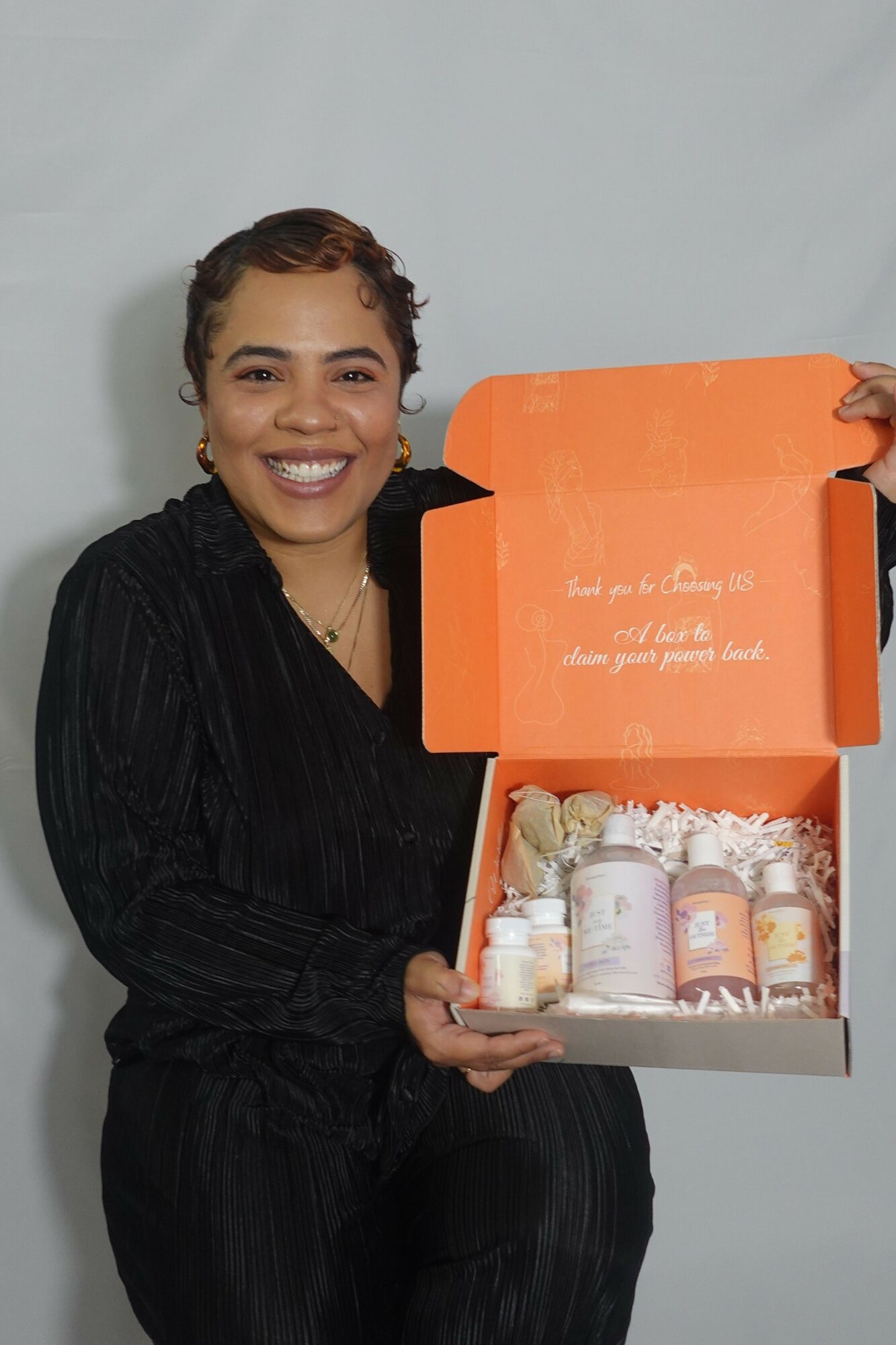 Woman smiling holding an open orange box with skincare products inside, against a plain background.