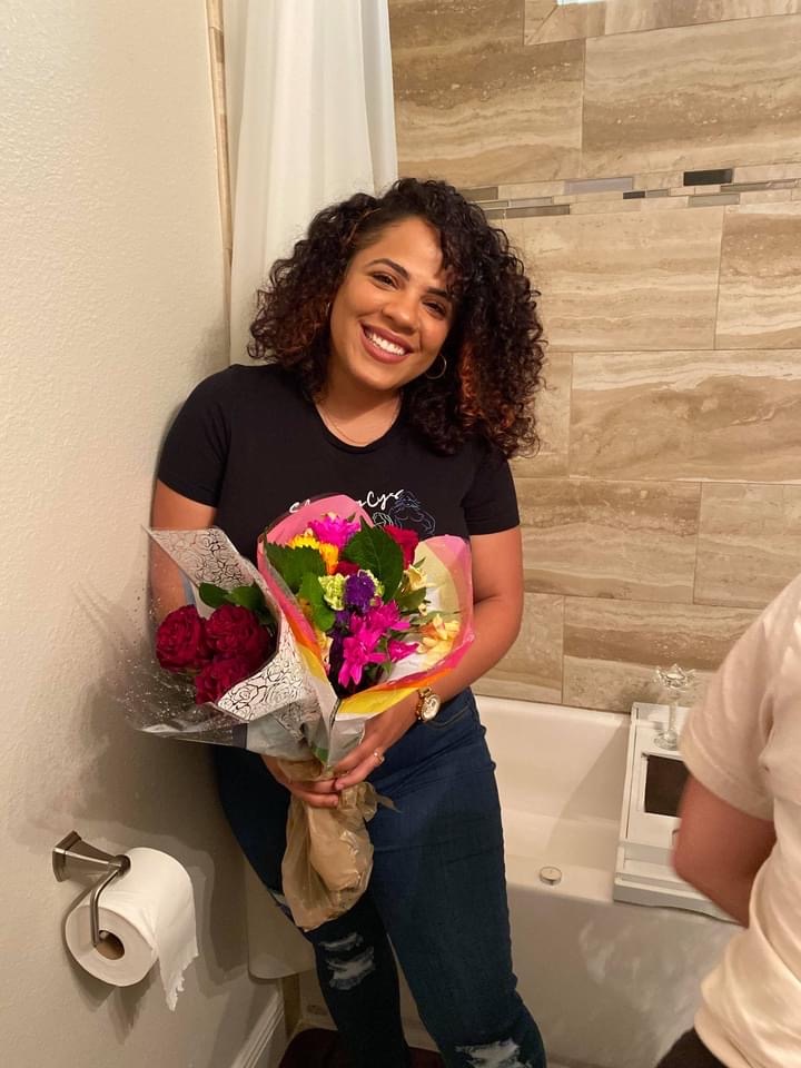 Smiling woman with curly hair holding a bouquet of colorful flowers in a bathroom.