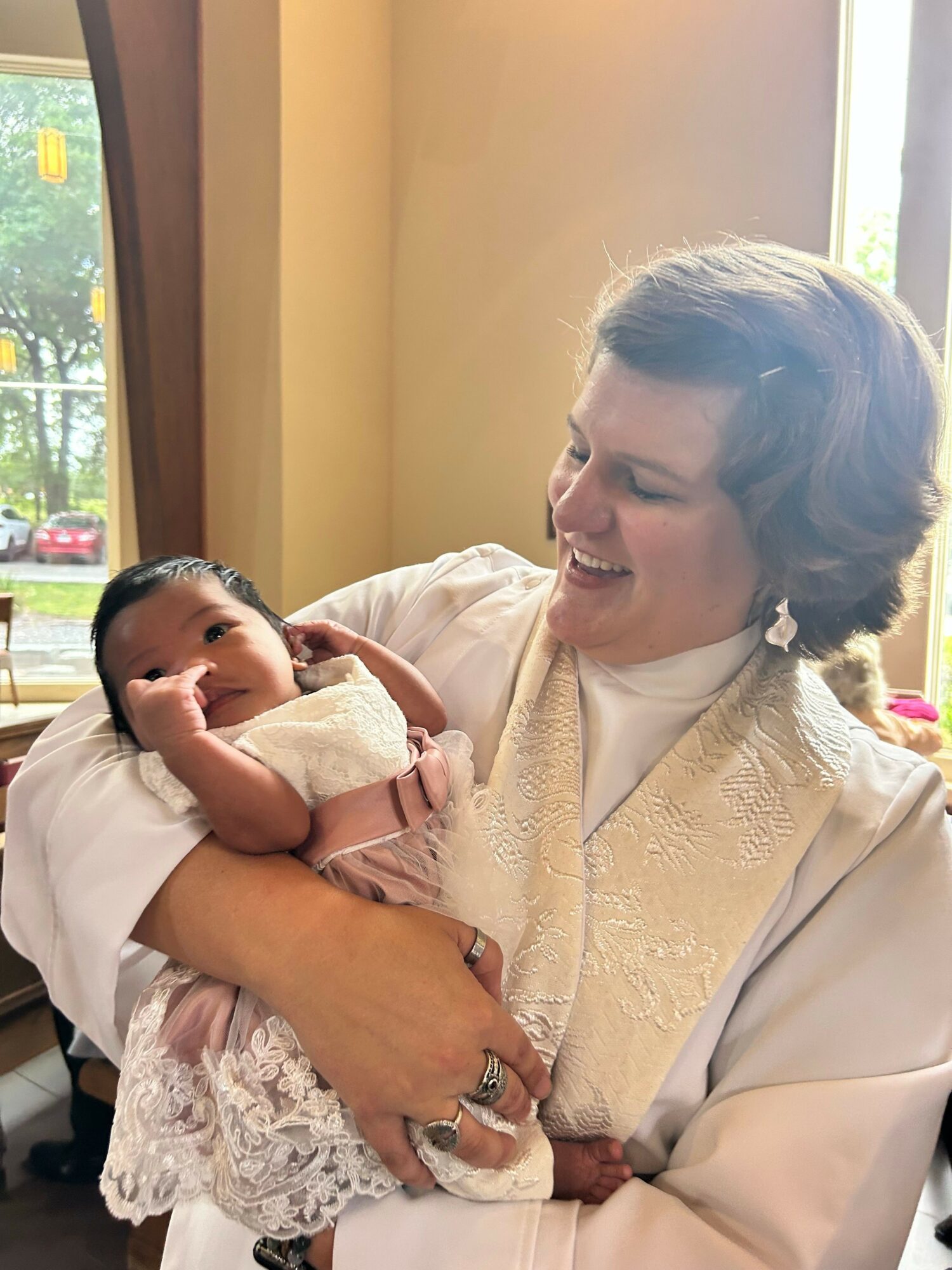 Woman holding a baby, smiling, indoors near a window with sunlight, woman wearing a light-colored top, baby in white clothing.
