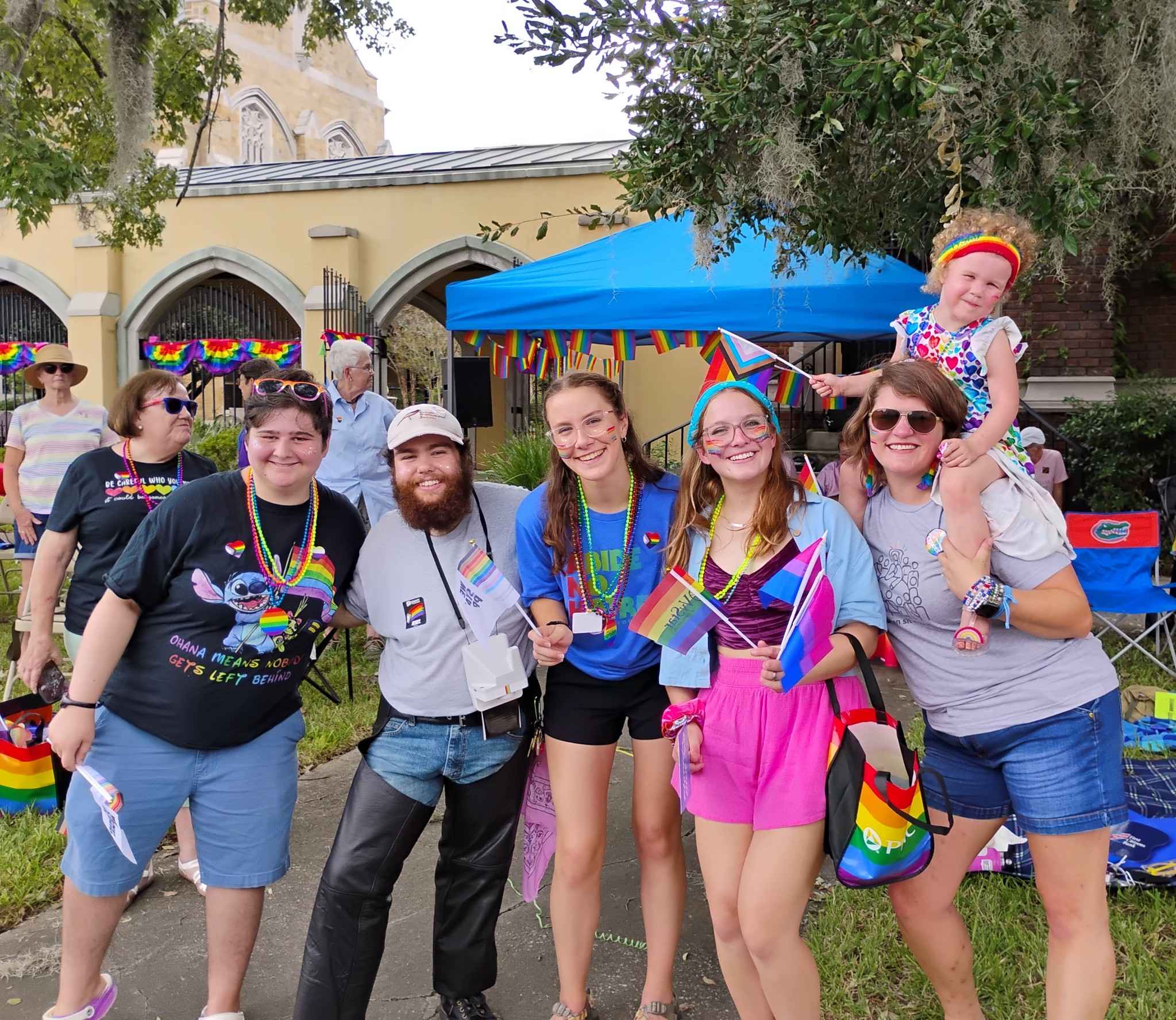 Group of people celebrating outdoors with rainbow decorations, some holding rainbow flags, and a child on a person's shoulders.