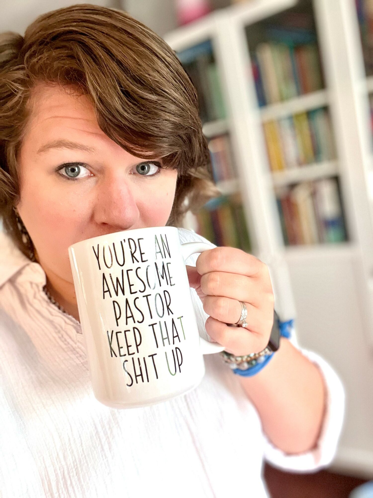 Person holding a mug with handwritten message, in front of bookshelves, with short brown hair and light skin, smiling.