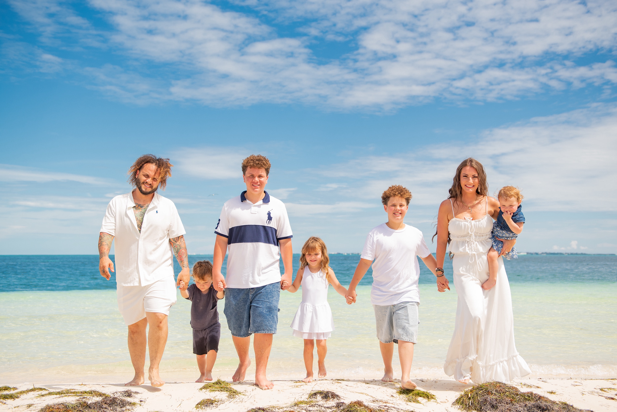 Group of six people holding hands on a beach, with blue sky and ocean in background.