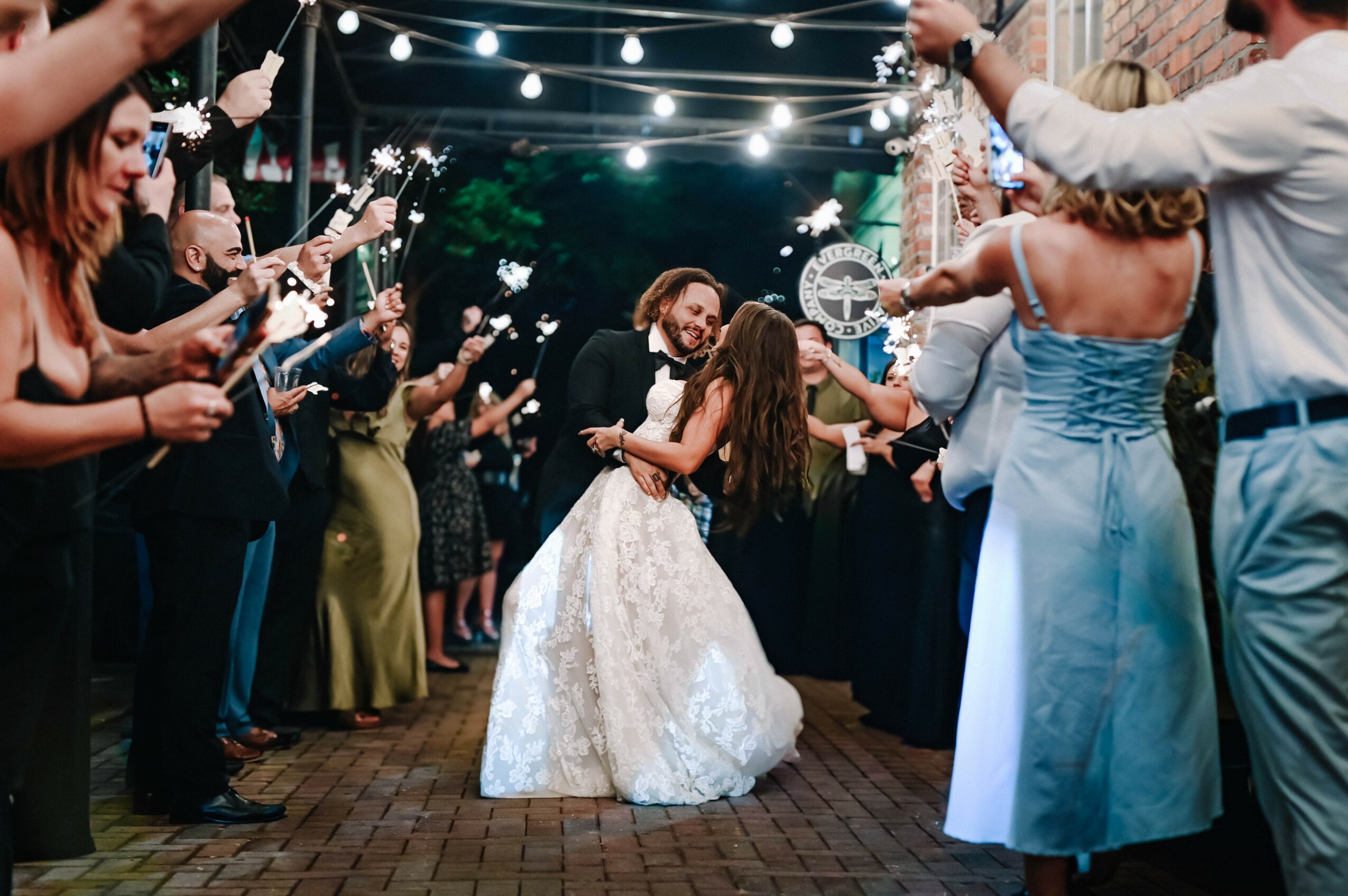 Couple dancing surrounded by guests at an outdoor celebration, string lights overhead, brick ground, evening setting.