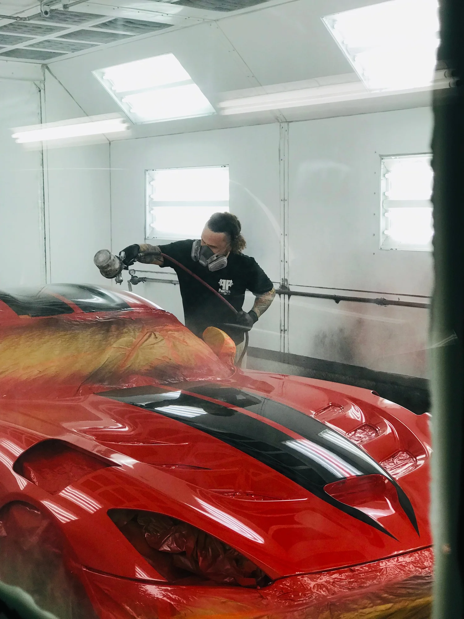 Person cleaning a red sports car inside a well-lit garage, wearing a black shirt and mask.