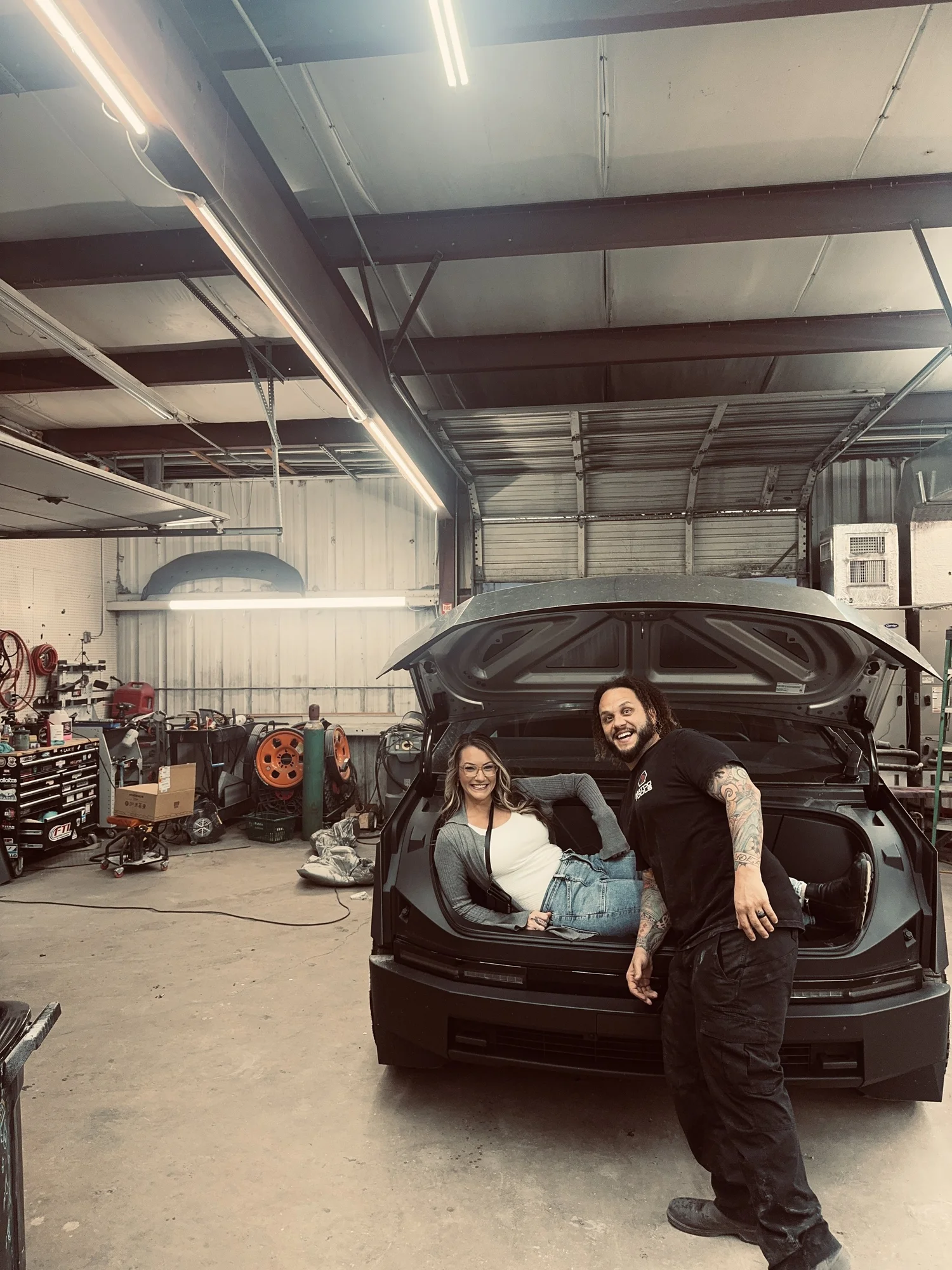 Two people sitting in the open trunk of a black car inside a garage, smiling at the camera.