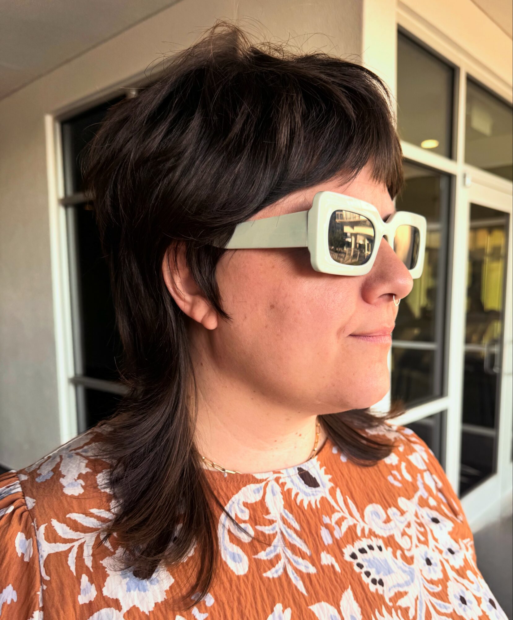 Woman with dark hair wearing large white sunglasses and an orange and white patterned top, standing outside near windows.