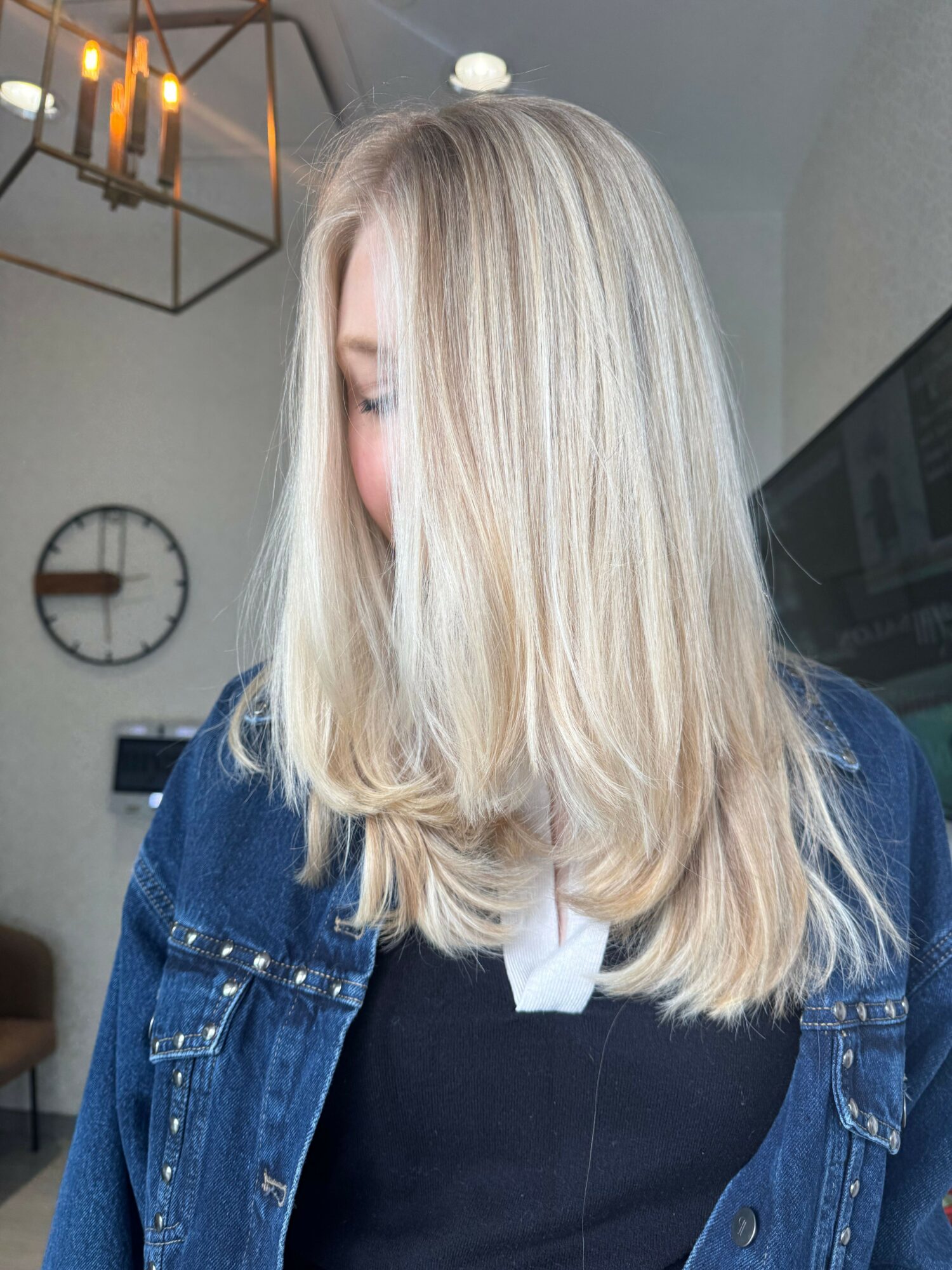 Person with long, blonde hair wearing a denim jacket, standing indoors with a clock and geometric light fixture in background.