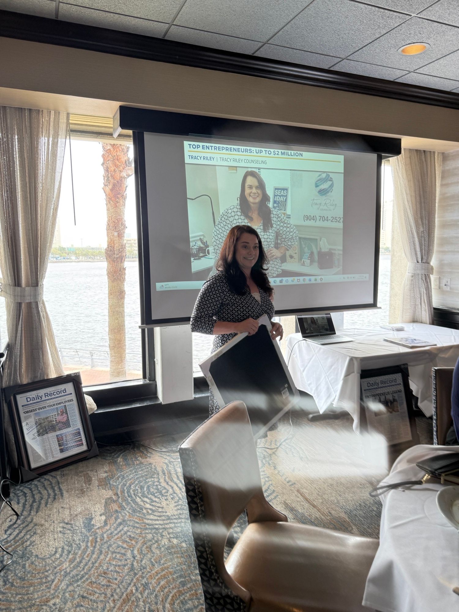 Woman standing in front of a presentation screen, speaking to an audience in a room with large windows and curtains.
