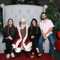 Four people sitting on a large, white, tufted chair with Christmas trees in the background, including a person dressed as Santa Claus.