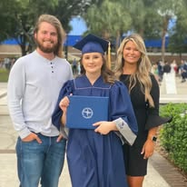 Three people outdoors, one in a graduation gown and cap holding a diploma, two others standing beside, smiling.
