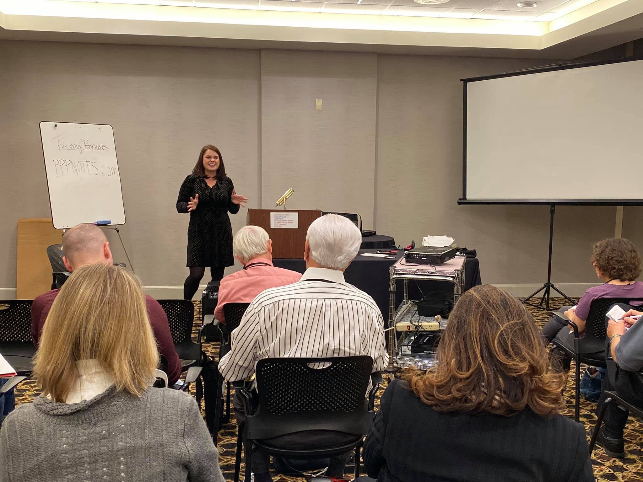 Woman standing and speaking in front of seated audience in a conference room, with a whiteboard and large screen behind her.