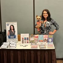 Woman holding a small dog standing behind a table with promotional materials, posters, and books, in an indoor setting.