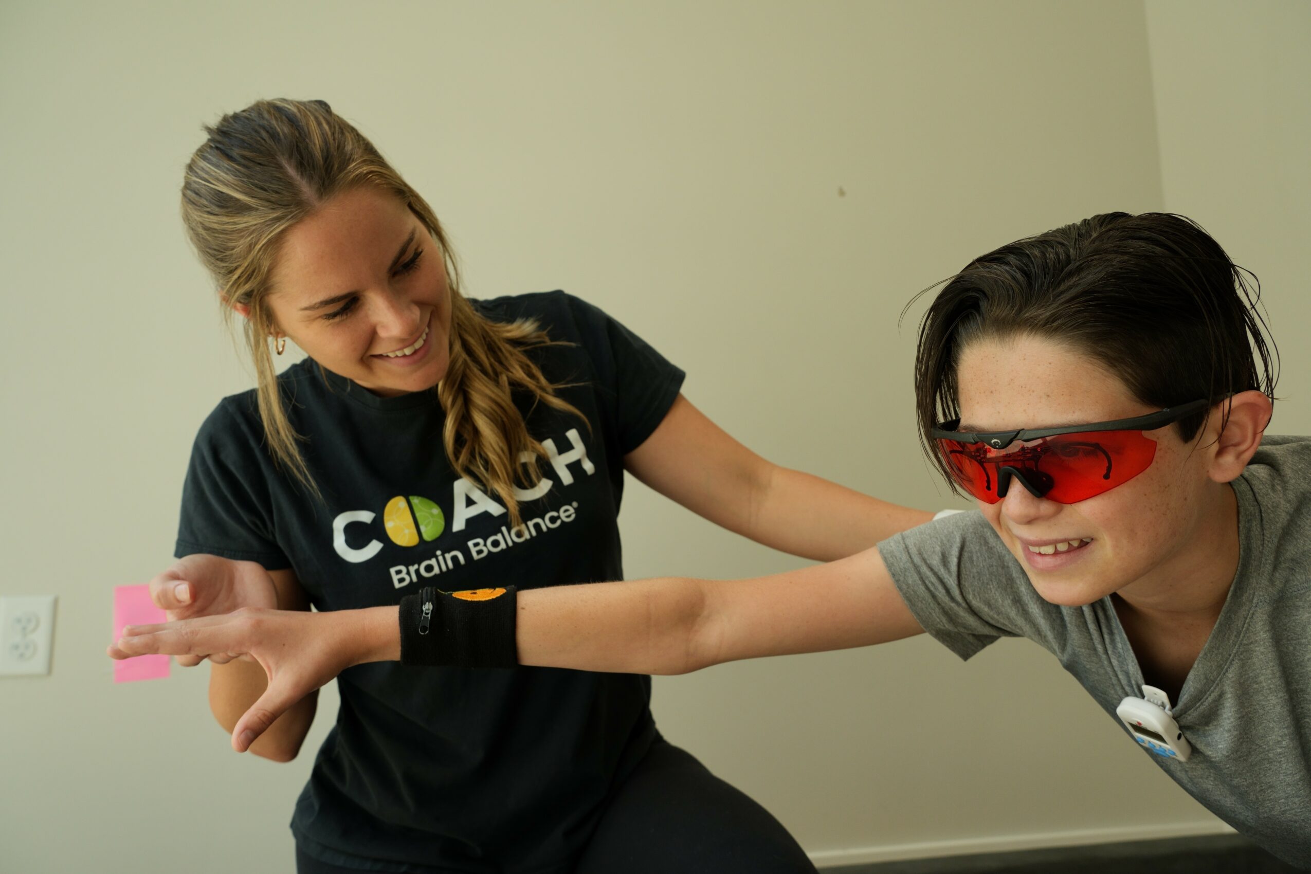 Two people smiling and playing with a toy, one wearing sunglasses, in a room with plain walls.