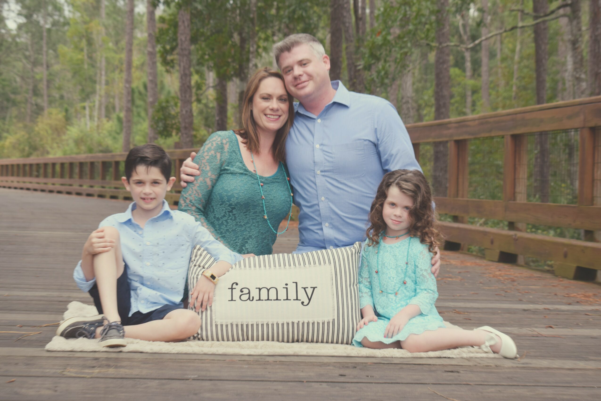 Family of four sitting on a blanket on a wooden bridge in a forest, with a sign reading 'family'.