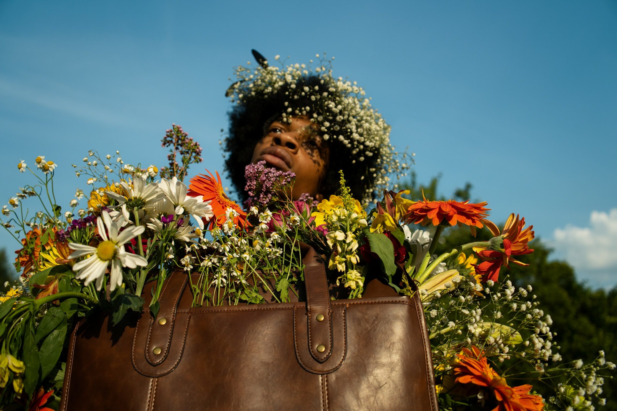 Person with curly hair smiling among colorful flowers in a large brown bag outdoors under a blue sky.